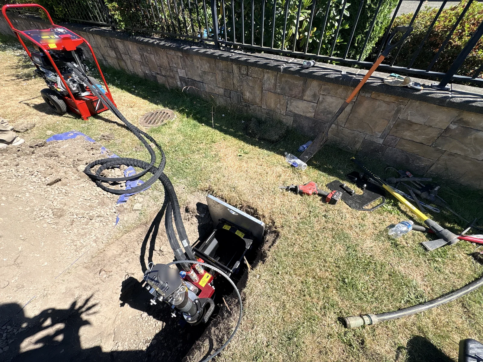 Bluetech trenchless rig on pull day: hydraulic power unit on the lawn feeding twin hydraulic lines to the pulling machine set in a small access pit